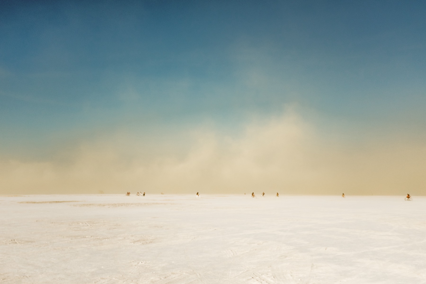 A pale, open salt plain under a deep blue sky, a band of dust haze
            along the horizon and a scattering of small distant figures crossing
            the flat.